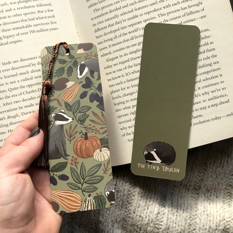 Op de afbeelding: Twee decoratieve bladwijzers. E&eacute;n heeft een das, pompoenen en bladeren op een groene achtergrond, met een bruine kwast. De andere is effen groen met een slapende das en de tekst "THE TINY TOUCAN".