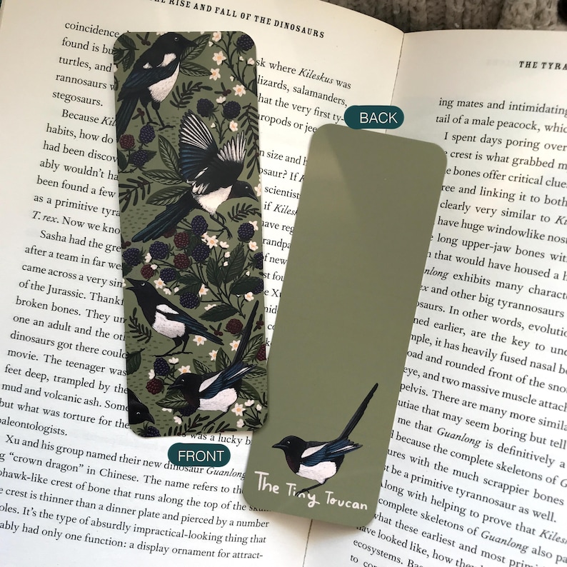 May include: Two rectangular bookmarks. One features a magpie bird design with berries and foliage on a dark green background. The other bookmark is solid olive green with a magpie illustration and the text "The Tiny Toucan".