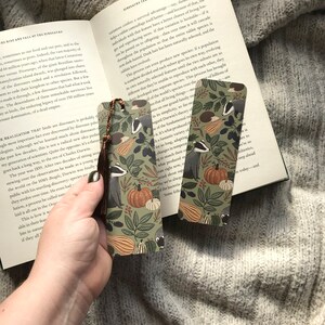 Op de afbeelding: Twee decoratieve bladwijzers met een herfstthema met dassen, pompoenen en bladeren in groen, oranje en bruin. Elke bladwijzer heeft een bruine kwast.