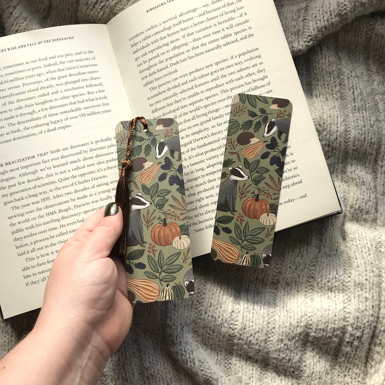 Op de afbeelding: Twee decoratieve bladwijzers met een herfstthema met dassen, pompoenen en bladeren in groen, oranje en bruin. Elke bladwijzer heeft een bruine kwast.