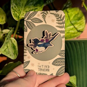 May include: A wooden pin depicting a blue, black, and white bird with purple berries. The pin is displayed on a card with a green circle and leaf design. The card is printed with the text "the tiny toucan".