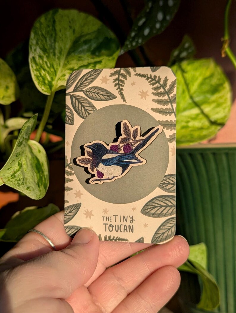 May include: A wooden pin depicting a blue, black, and white bird with purple berries. The pin is displayed on a card with a green circle and leaf design. The card is printed with the text "the tiny toucan".
