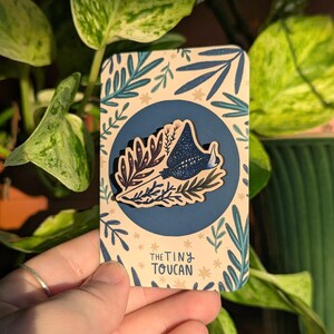 May include: A toucan-themed pin set on a card with a botanical design. The pin has a navy blue background with a starry pattern. The card has a light beige background with blue and green leaves and the text "THE TINY TOUCAN".