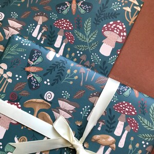 May include: Gift wrap featuring a whimsical forest design with mushrooms, leaves, and insects in shades of teal, brown, and white. A cream-colored ribbon is tied around the wrapped package. The overall theme is nature and woodland.