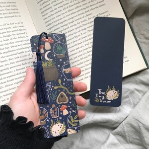 May include: Two navy blue bookmarks. One has a dark blue tassel and a pattern of mushrooms, plants, and crystals. The other is solid navy blue with the text "The Tiny Toucan" and a skull graphic.