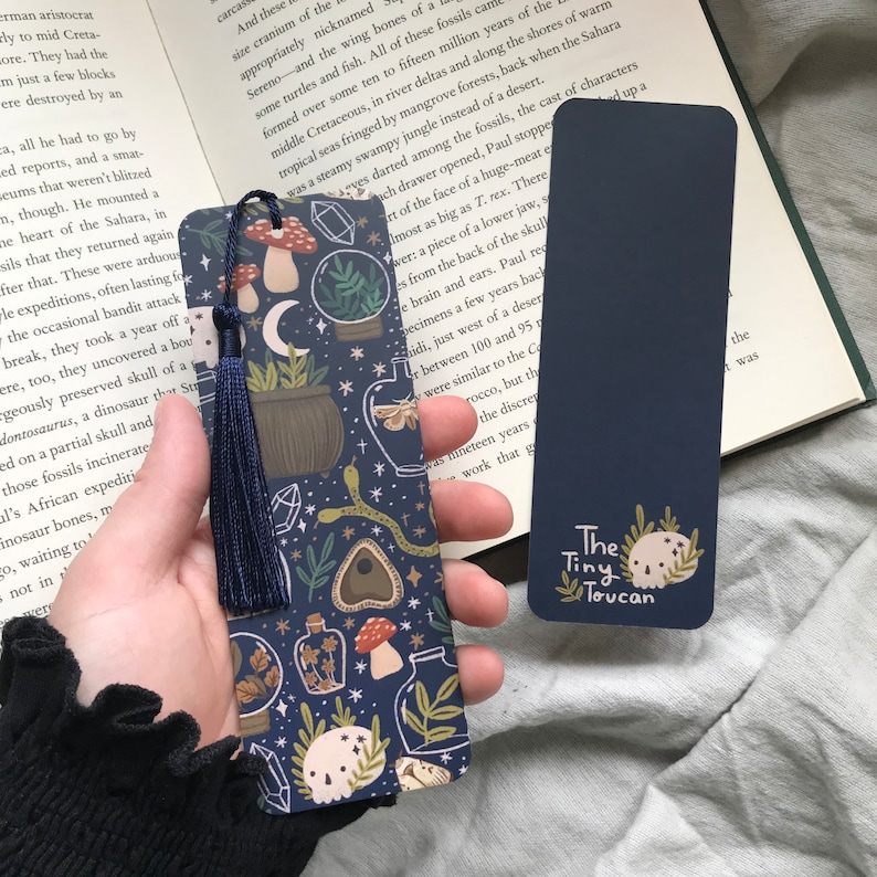 May include: Two navy blue bookmarks. One has a dark blue tassel and a pattern of mushrooms, plants, and crystals. The other is solid navy blue with the text "The Tiny Toucan" and a skull graphic.