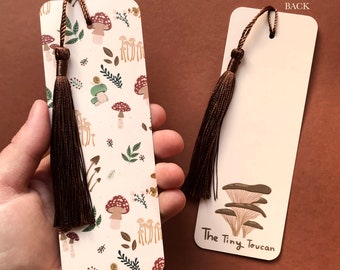 May include: A white bookmark with a brown tassel. The front of the bookmark features a pattern of mushrooms and leaves. The back of the bookmark has the text "The Tiny Toucan".