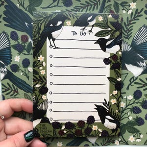 May include: A rectangular notepad with a green background featuring a pattern of magpies, berries, and flowers. The notepad has a "To Do" header and lined spaces. The bottom left corner has the text "THE TINY TOUCAN".