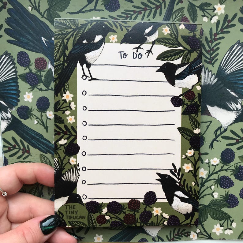May include: A rectangular notepad with a green background featuring a pattern of magpies, berries, and flowers. The notepad has a "To Do" header and lined spaces. The bottom left corner has the text "THE TINY TOUCAN".