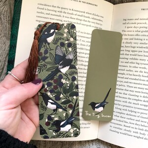 May include: Two decorative bookmarks featuring a bird and berry design. One is olive green with a brown tassel, showing illustrations of black and white birds and berries. The other is a plain olive green bookmark with a single bird illustration. The text "The Tiny Toucan" is visible.