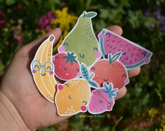 May include: A hand holding a set of colorful fruit stickers. The stickers feature a banana, pear, watermelon, tomato, orange, lemon, and strawberry. Each fruit has a cute cartoon face and some have glasses.