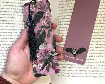 Puede incluir: Dos marcapáginas decorativos. Uno presenta un patrón repetido de murciélagos, flores y follaje sobre un fondo malva, con una borla azul marino. El otro es de color malva liso con un gráfico de murciélago y el texto "THE TINY TOUCAN".