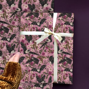 May include: Gift wrap featuring a repeating pattern of black bats, white flowers, and berries on a mauve background. A cream-colored ribbon is tied around the wrapped gift. The overall theme is a whimsical, gothic aesthetic.