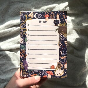 May include: A notepad with a dark blue background and a decorative border featuring mystical illustrations. The notepad has the words "TO DO" at the top and lined spaces for writing. The border includes images of mushrooms, potion bottles, and moons.
