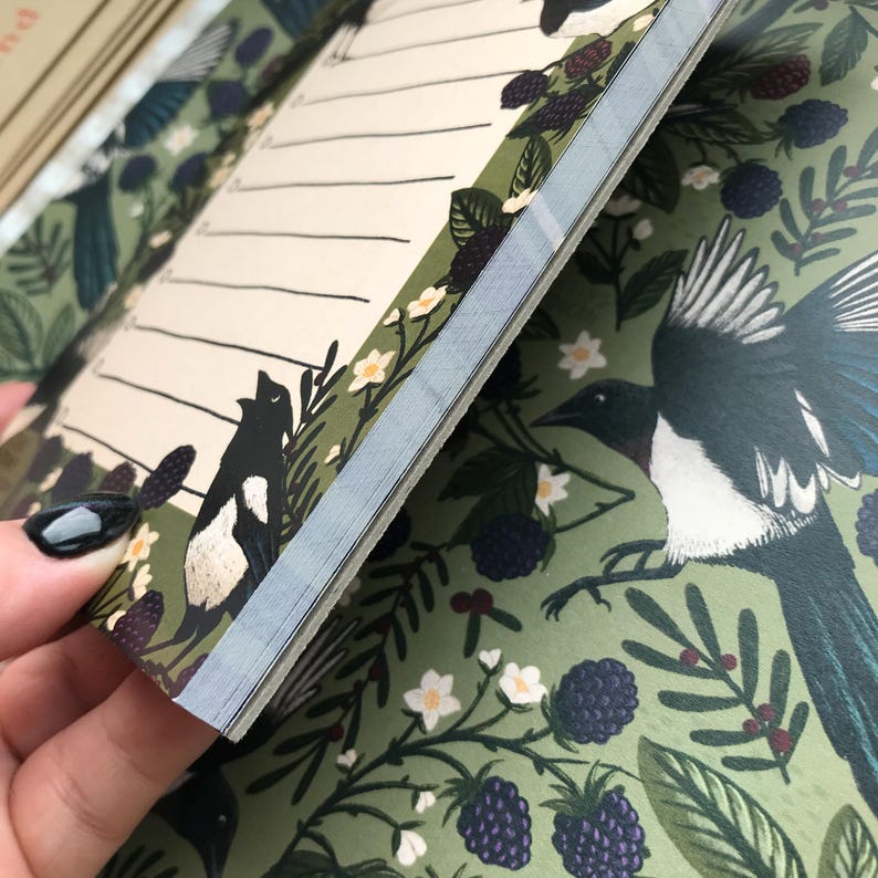 May include: Open journal with a green cover featuring a magpie illustration, white flowers, and blackberries. The interior pages have lined spaces for writing. The journal's edges are light blue.