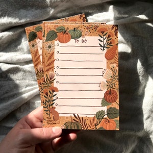 May include: A stack of to-do list notepads with an autumnal theme. The paper is lined and has a border of pumpkins, leaves, and flowers in shades of orange, brown, and green. The text "To Do" is printed at the top of the notepad. The bottom of the notepad has the text "The Tiny Toucan".