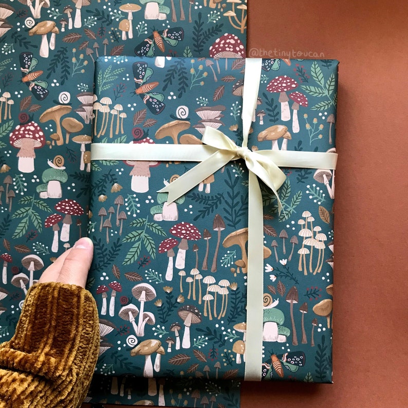 May include: Gift-wrapped package featuring a teal background with a whimsical mushroom and foliage pattern. The package is tied with a cream-colored ribbon. The design includes various mushrooms, leaves, and small insects. The overall aesthetic is nature-inspired and decorative.
