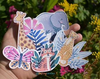 May include: A set of five colourful animal stickers featuring a giraffe, elephant, cheetah, tiger, and butterfly. The stickers are surrounded by blue and green leaves and pink flowers. The text "@thetinytoucan" is visible at the bottom of the image.