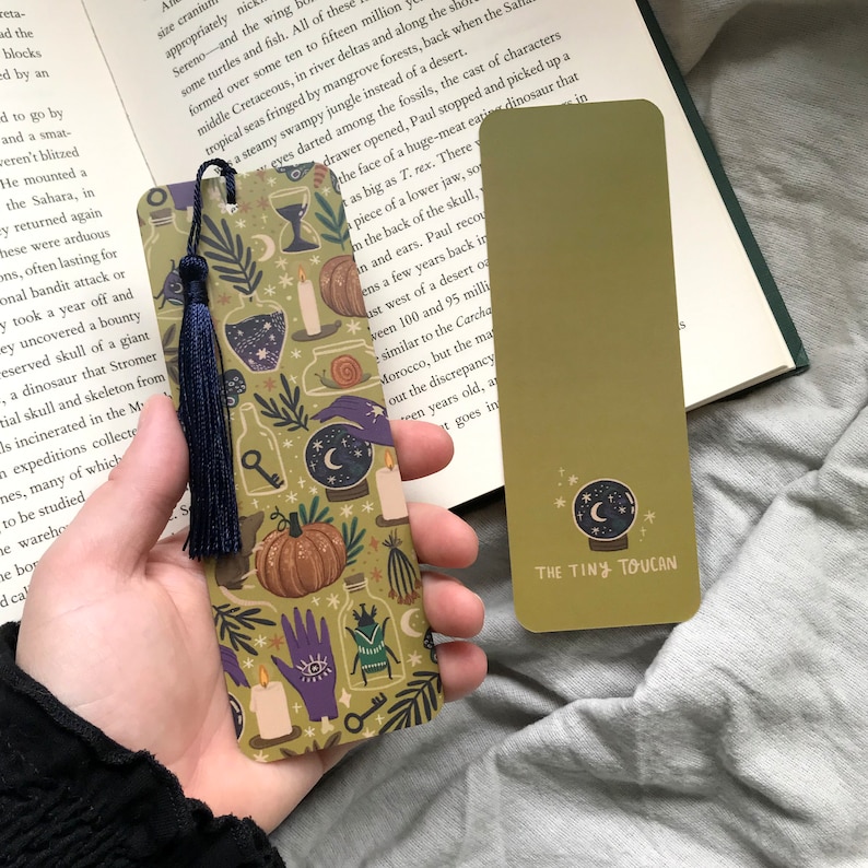 May include: Two decorative bookmarks. One has a navy tassel and a pattern of pumpkins, potion bottles, and a crystal ball on a green background. The other is plain green with a crystal ball graphic and the text "THE TINY TOUCAN".