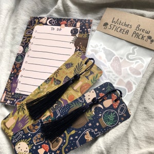 May include: A selection of witch-themed stationery. The image shows a notepad with "To Do" text, two bookmarks with black tassels, and a sticker pack. The items feature illustrations of mushrooms, moons, and potion bottles.