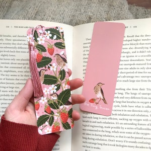 May include: A pink bookmark with a floral pattern featuring strawberries, white flowers, and two brown birds. The bookmark has a pink tassel. The text "The Tiny Toucan" is printed on the bookmark.