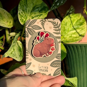 Op de afbeelding: Een rechthoekige sticker met een bruine capibara, rode bloemen en groene bladeren. De sticker heeft een lichtbeige achtergrond met een bloemenontwerp en de tekst "THE TINY TOUCAN".