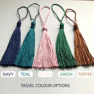 Op de afbeelding: Vijf kleurrijke kwasten in marineblauw, blauwgroen, roze, groen en toffee. Elke kwast heeft een lus en een geknoopte basis. De tekst "Tassel Colour Options" wordt eronder weergegeven, met de kleurnamen.