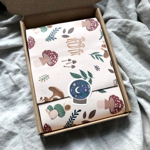 May include: An open cardboard box with a package inside, wrapped in cream paper with a mushroom and foliage design. A sticker of a crystal ball with a crescent moon and stars is on the wrapping. The box is on a grey surface.