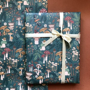 May include: Two rolls of gift wrap with a dark teal background and a repeating pattern of mushrooms, leaves, snails, and butterflies. One roll is wrapped with a cream-colored ribbon tied in a bow.