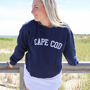 May include: A navy blue sweatshirt with the text "CAPE COD" in white lettering. The sweatshirt is worn by a person standing on a wooden boardwalk.