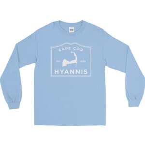 May include: Light blue long-sleeved t-shirt with a white graphic of a vintage-style stamp featuring the outline of Cape Cod, the text "Cape Cod", "Inc", "1639", and "Hyannis".