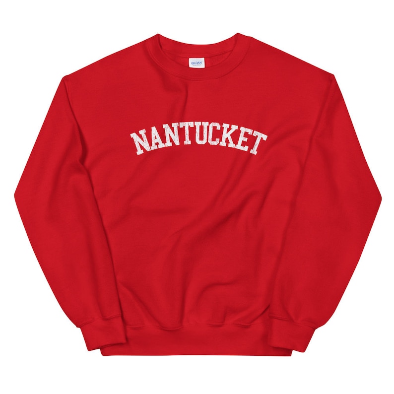 May include: A red sweatshirt with the word "NANTUCKET" printed across the chest in white, arched lettering. The sweatshirt has a crew neck and long sleeves. The Gildan brand label is visible at the back of the neck.