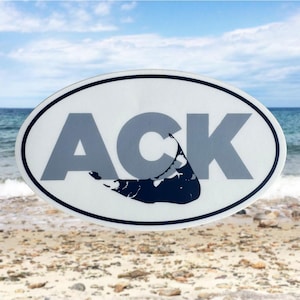 May include: Oval sticker with the letters "ACK" in gray, with a dark blue outline of a landmass. The sticker has a white background and a black border. The background shows a beach and ocean.
