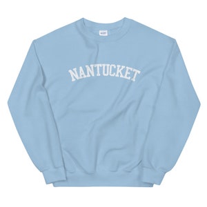 May include: A light blue sweatshirt with the word "NANTUCKET" printed across the chest in white, arched lettering. The sweatshirt is a crew neck style and appears to be made of a soft material. The Gildan logo is visible on the upper back.