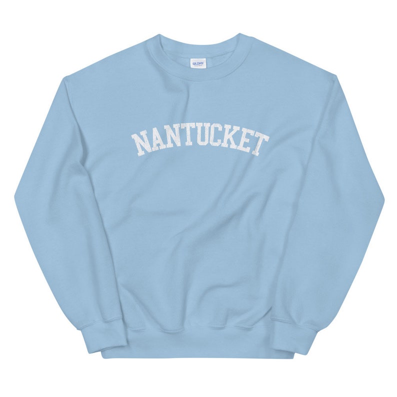 May include: A light blue sweatshirt with the word "NANTUCKET" printed across the chest in white, arched lettering. The sweatshirt is a crew neck style and appears to be made of a soft material. The Gildan logo is visible on the upper back.