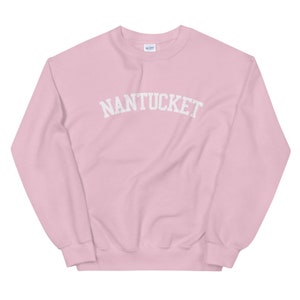 May include: A light pink sweatshirt with the word "NANTUCKET" arched across the chest in white block letters. The sweatshirt has a crew neck and long sleeves. The Gildan logo is visible on the upper back.