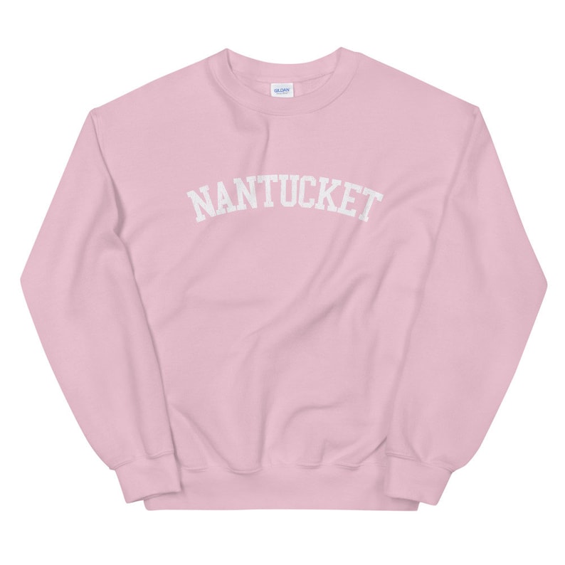May include: A light pink sweatshirt with the word "NANTUCKET" arched across the chest in white block letters. The sweatshirt has a crew neck and long sleeves. The Gildan logo is visible on the upper back.