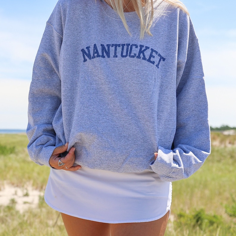 May include: A gray sweatshirt with the word "NANTUCKET" in navy blue arched across the chest. The person is wearing a white skirt. The background shows a beach and blue sky.