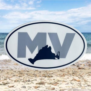 May include: Oval sticker with a white background, black border, and gray letters "MV". A dark blue silhouette of an island is at the bottom. The sticker is set against a beach and ocean backdrop.