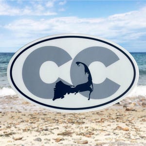 May include: Oval sticker with a white background, featuring two gray "CC" letters and a dark blue silhouette of a peninsula. The sticker has a black border and is set against a beach and ocean backdrop.