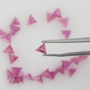 Könnte beinhalten: Nahaufnahme eines rosafarbenen dreieckigen Edelsteins, der mit einer Pinzette gehalten wird. Der Edelstein ist von einer Streuung ähnlicher rosafarbener dreieckiger Edelsteine auf einer weißen Oberfläche umgeben. Der Text "GemsRainbowCo" ist in der unteren rechten Ecke des Bildes sichtbar.