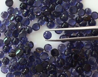 May include: A large group of round, dark blue gemstones, some with a slight iridescent sheen, are scattered on a white surface. A pair of tweezers holds one of the gemstones.