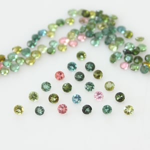 May include: Assortment of round, faceted gemstones in various shades of green, blue, and pink. The gemstones are scattered on a white surface.
