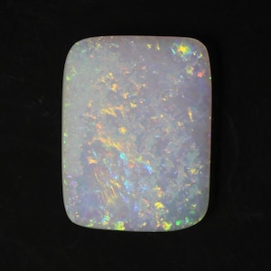 May include: A rectangular, polished opal gemstone with rounded corners. The opal displays a milky white base color with flashes of iridescent colors, including yellow, green, and blue. The gemstone is set against a black background.