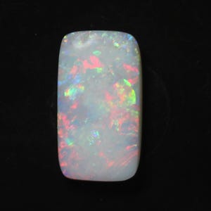 Könnte beinhalten: Ein rechteckiger, polierter Opal-Edelstein mit weißer Basis und lebhaftem internen Farbspiel. Der Edelstein zeigt Blitze von Rot, Grün und Blau vor dunklem Hintergrund. Die Oberfläche des Opals ist glatt und abgerundet.