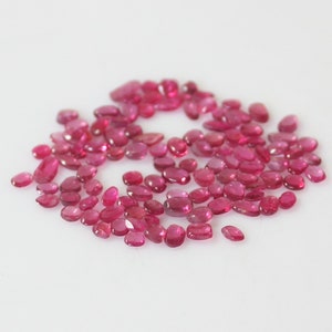 May include: A collection of loose pink ruby gemstones, oval shaped, scattered on a white surface.