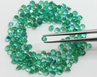 May include: A close-up of a small, round, green emerald gemstone held by tweezers. The emerald is surrounded by a circle of many other loose emeralds.