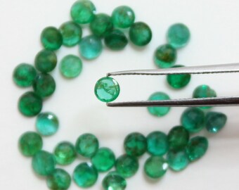 May include: A scattering of small, round, emerald-green gemstones on a white background. One gem is held by tweezers. The gems vary in shade, with some appearing more translucent. The image is well-lit, highlighting the gems' colour and clarity.