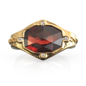 Könnte beinhalten: Ein Goldring mit einem großen, facettierten, sechseckigen, tiefroten Granat-Edelstein in der Mitte. Die Ringschiene besteht aus poliertem Gelbgold und ist mit vier kleinen, runden, klaren weißen Diamanten an den Eckpunkten besetzt.