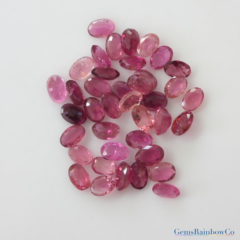 May include: A collection of pink oval-shaped gemstones. The gemstones are various shades of pink, from light to dark. The gemstones are arranged in a random pattern on a white surface.
